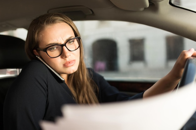 Joven empresaria seria con vestido negro y anteojos hablando por teléfono celular mirando cuidadosamente los documentos mientras se sienta detrás del volante en el auto