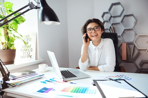 Joven empresaria sonriente hermosa hablando por teléfono en el lugar de trabajo en la oficina.