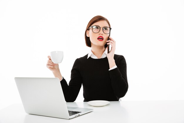 Joven empresaria con una taza de café o té y una computadora portátil, hablando por teléfono