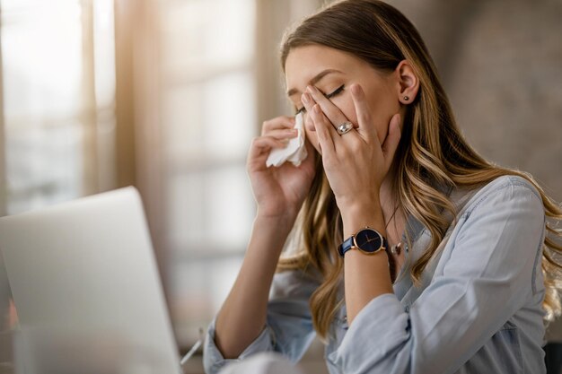 Joven empresaria con virus del resfriado y la gripe que tiene dolor de cabeza mientras trabaja en la oficina