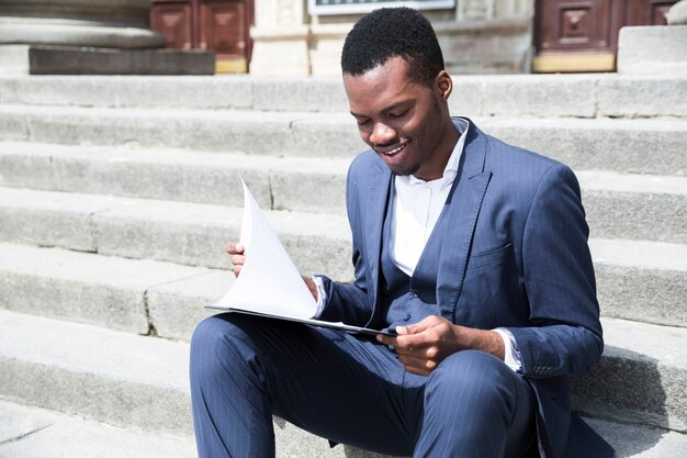 Un joven empresario africano hablando por teléfono móvil sentado en la escalera con la computadora portátil