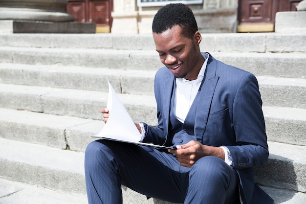 Un joven empresario africano hablando por teléfono móvil sentado en la escalera con la computadora portátil