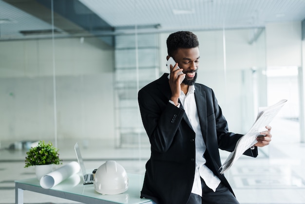 Joven empresario afroamericano leyendo el periódico y hablando por teléfono en su oficina