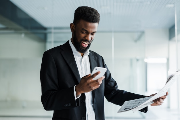 Joven empresario afroamericano leyendo el periódico y hablando por teléfono en su oficina