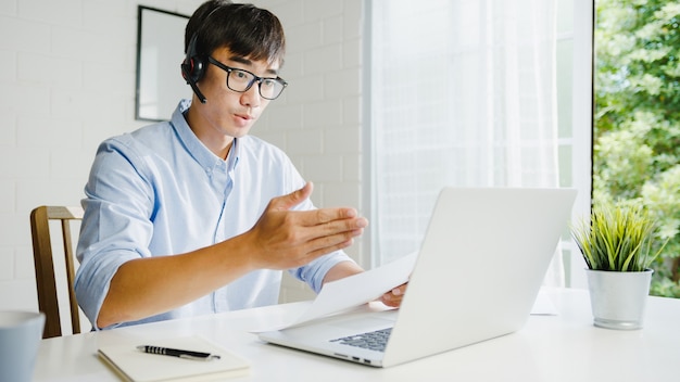 Joven empresario de Asia usando una computadora portátil hablar con sus colegas sobre el plan en la videollamada mientras trabaja de manera inteligente desde casa en la sala de estar Autoaislamiento, distanciamiento social, cuarentena para la prevención del coronavirus.