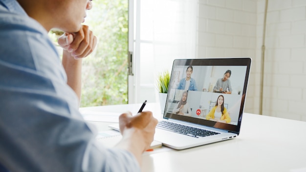 Foto gratuita joven empresario de asia usando laptop hablar con colegas sobre el plan en la reunión de videollamada mientras trabaja desde casa en la sala de estar. autoaislamiento, distanciamiento social, cuarentena para la prevención del coronavirus.