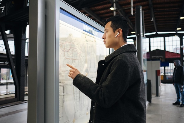 Joven empresario asiático con auriculares inalámbricos mirando con confianza la ruta del transporte público en la estación de metro