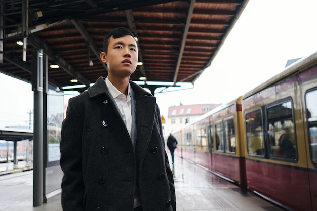 Foto gratuita joven empresario asiático con estilo mirando atentamente hacia otro lado mientras espera el tren en la plataforma del metro