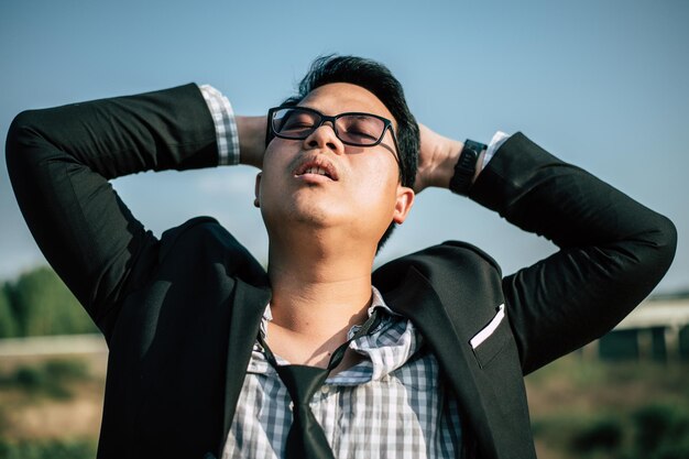 Joven empresario asiático vestido con traje con corbata y anteojos cierra los ojos con cansancio y duerme para relajarse al aire libre