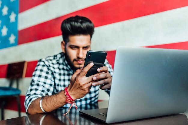 Joven empresario de barba negra en ropa casual está usando un teléfono inteligente y sonriendo mientras trabaja con una computadora portátil en un gastropub.