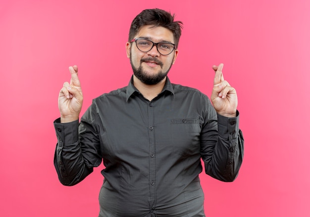 Joven empresario complacido con gafas y cruzando los dedos aislado en la pared rosa