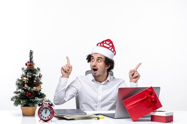 Joven empresario confundido con sombrero de santa claus divertido apuntando algo en diferentes direcciones en la oficina sobre fondo blanco.