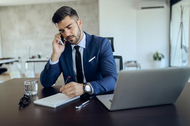 Joven empresario escribiendo notas mientras habla por teléfono en la oficina