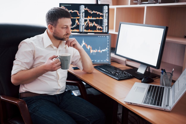 Joven empresario está sentado en la oficina en la mesa, trabajando en equipo con muchos monitores, diagramas en monitor. El corredor de bolsa analiza gráficos de opciones binarias. Hombre hipster tomando café, estudiando