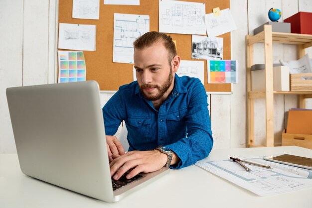 Joven empresario exitoso sonriendo, sentado en el lugar de trabajo escribiendo en la computadora portátil