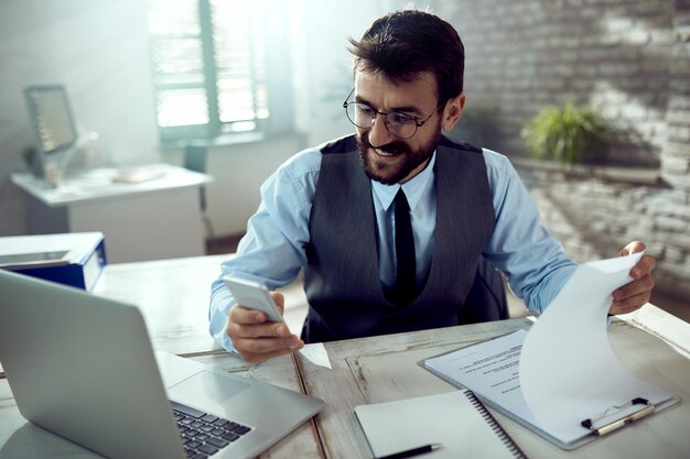 Joven empresario feliz revisando informes y usando un teléfono inteligente en la oficina