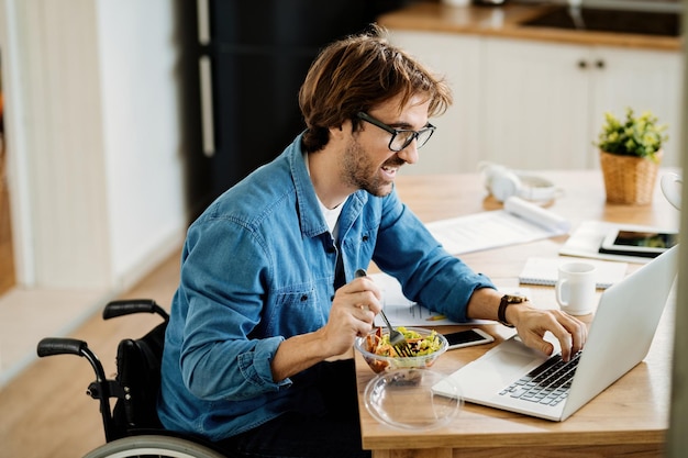 Joven empresario feliz en silla de ruedas navegando por la red en una computadora portátil mientras come saludable en casa