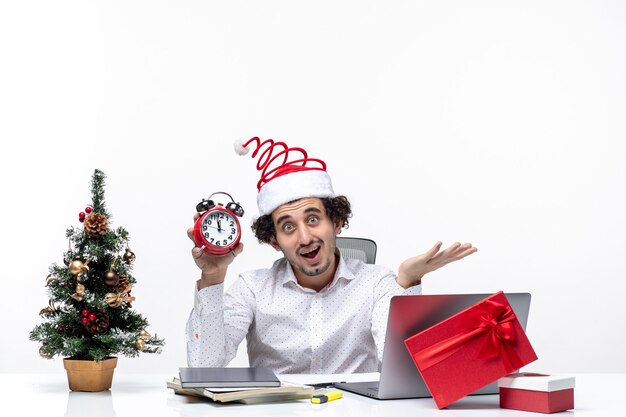 Joven empresario feliz con sombrero de santa claus y sosteniendo el reloj y apuntando algo en la oficina sobre fondo blanco.