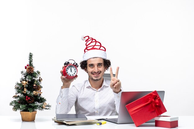 Joven empresario feliz con sombrero de santa claus y sosteniendo el reloj y mostrando dos en la oficina sobre fondo blanco.