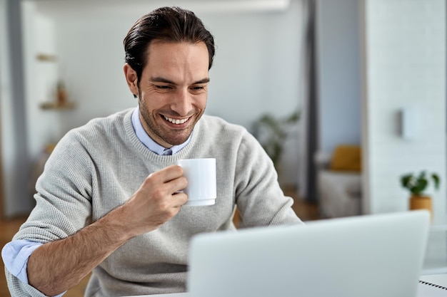 Joven empresario feliz trabajando en una computadora mientras bebe café en casa