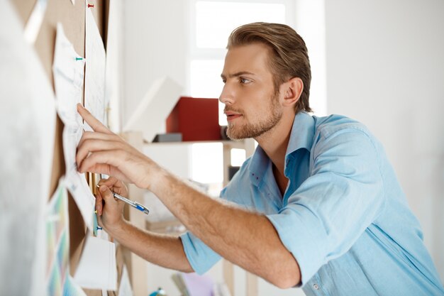 Joven empresario guapo señalar con dedo y escribir en el papel clavado en el panel de corcho.