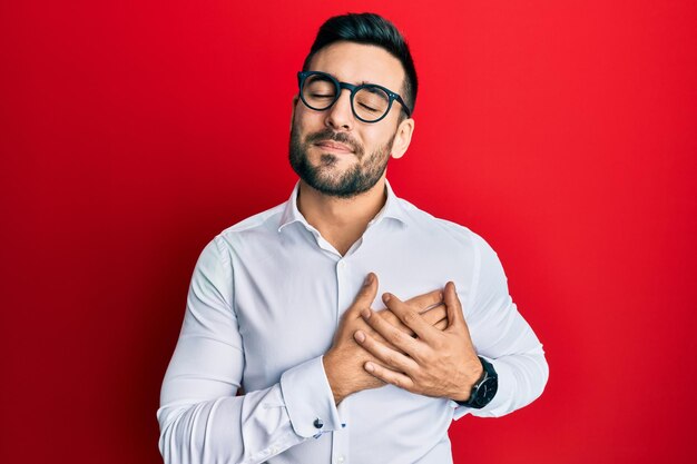 Joven empresario hispano con camisa y gafas sonriendo con las manos en el pecho con los ojos cerrados y un gesto agradecido en la cara. concepto de salud