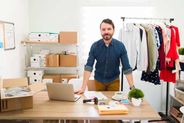 Joven empresario hispano detrás de su escritorio de oficina y trabajando en hacer paquetes con ropa de moda para enviar a sus clientes