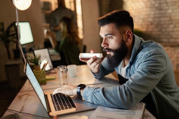 Joven empresario leyendo un correo electrónico en la computadora mientras graba un mensaje de voz en un teléfono inteligente durante el trabajo nocturno en la oficina