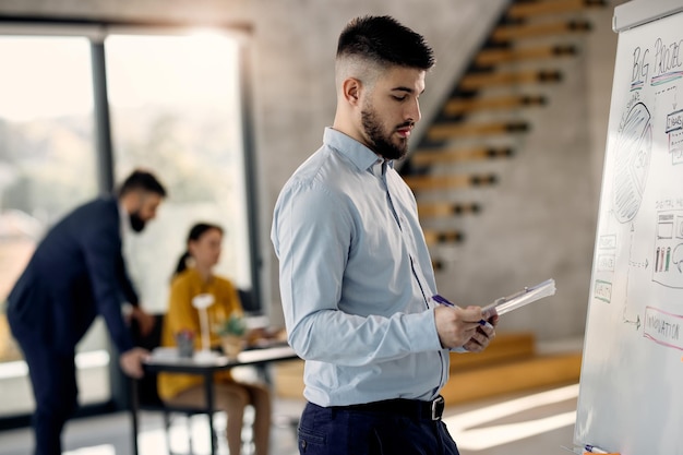 Joven empresario leyendo notas mientras dibuja un plan en la pizarra en la oficina