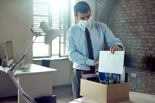 Joven empresario con mascarilla y empacando sus pertenencias después de perder su trabajo debido a la pandemia de COVID19