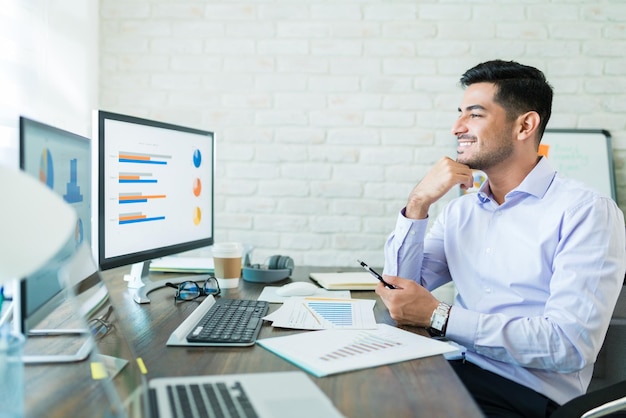 Joven empresario mirando gráficos en la computadora mientras está sentado con el teléfono inteligente