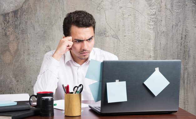 Joven empresario mirando portátil con expresión enojada en el escritorio de oficina.