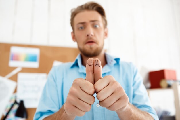 Joven empresario molesto guapo mostrando los pulgares para arriba con dibujos de caras divertidas. Centrarse en las manos. Interior de oficina moderno blanco