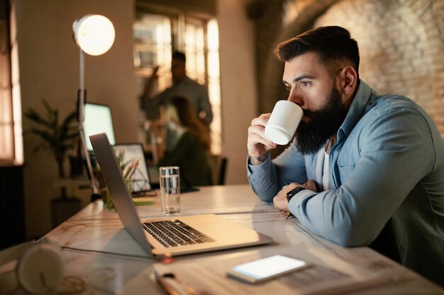 Joven empresario navegando por la red en una computadora mientras bebe café por la noche en la oficina