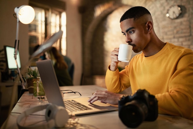 Joven empresario negro bebiendo café mientras trabaja hasta tarde en una computadora en la oficina