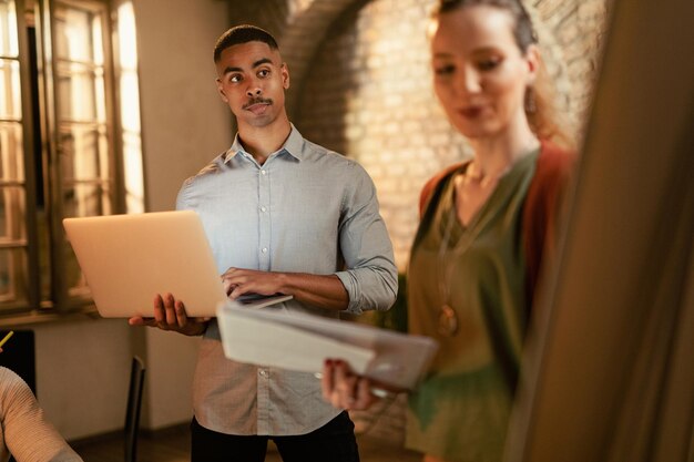 Foto gratuita joven empresario negro que usa una computadora portátil mientras trabaja en nuevas ideas con sus colegas durante la presentación en la oficina