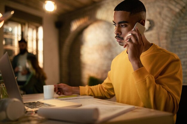 Joven empresario negro trabajando hasta tarde en una computadora y comunicándose por teléfono celular en la oficina
