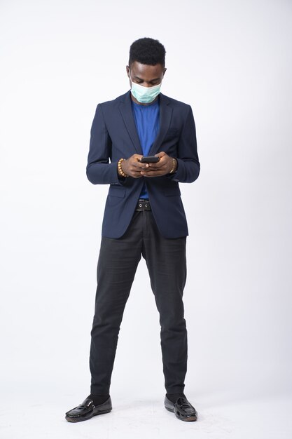Joven empresario negro vistiendo un traje y mascarilla usando su teléfono delante de un blanco