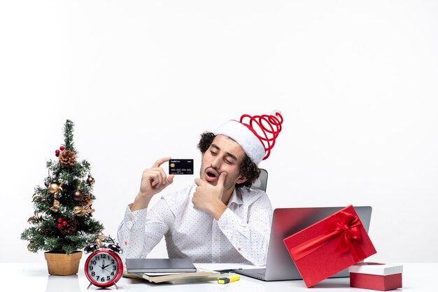 Joven empresario ocupado con sombrero de santa claus y sosteniendo su tarjeta bancaria intercambiando ideas cuidadosamente en la oficina sobre fondo blanco.