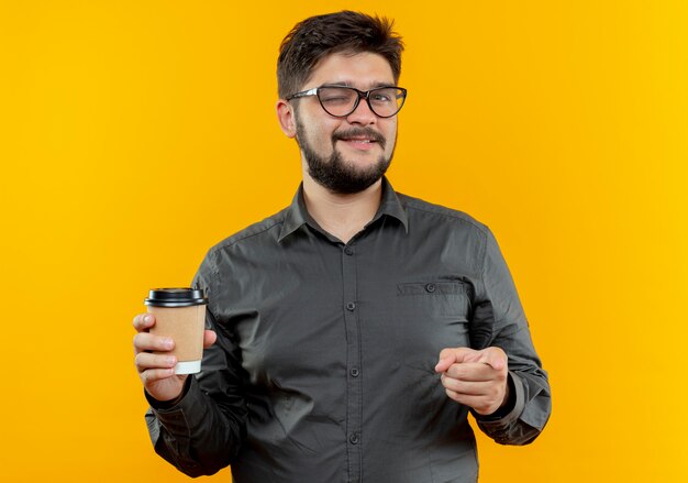 Joven empresario parpadeó con gafas sosteniendo una taza de café y mostrando gesto aislado sobre fondo amarillo