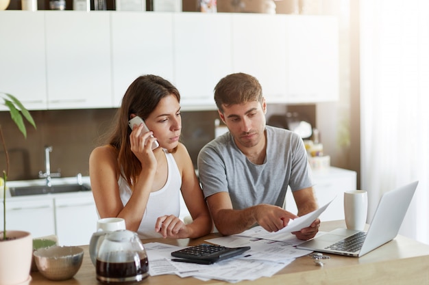 Joven empresario señalando con el dedo al documento, tratando de expandir algo a su esposa que está hablando por teléfono inteligente. Pareja revisando sus cuentas bancarias y calculando cifras anuales