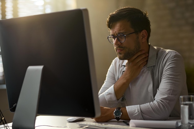 Joven empresario siente dolor en la garganta mientras trabaja en una PC de escritorio en la oficina