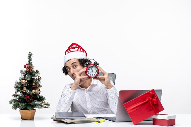 Joven empresario con sombrero de santa claus y mostrando el reloj mirando algo con sorpresa y sentado en la oficina sobre fondo blanco.