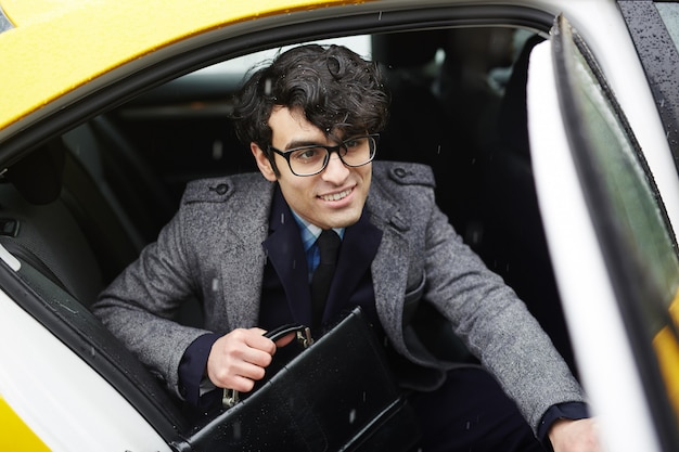 Joven empresario sonriente dejando taxi bajo la lluvia