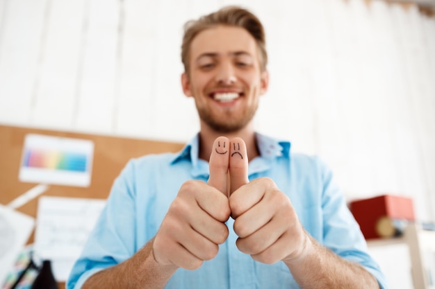 Foto gratuita joven empresario sonriente guapo mostrando los pulgares para arriba con dibujos de caras divertidas. centrarse en las manos. interior de oficina moderno blanco