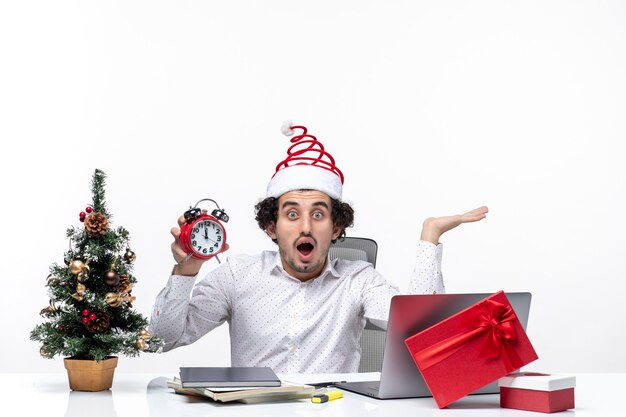 Joven empresario sorprendido con sombrero de santa claus y mostrando el reloj y sentado en la oficina sobre fondo blanco.