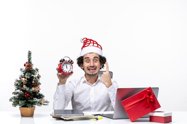 Joven empresario sorprendido con sombrero de santa claus y sosteniendo el reloj y mostrando uno en la oficina sobre fondo blanco.