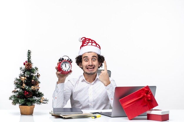 Joven empresario sorprendido con sombrero de santa claus y sosteniendo el reloj y mostrando uno en la oficina sobre fondo blanco.