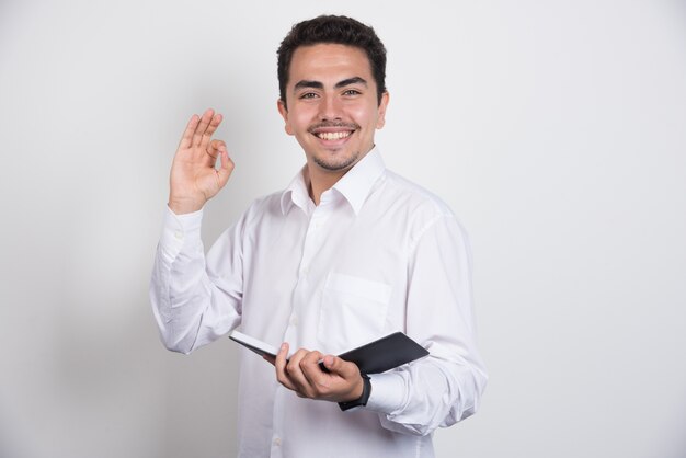 Joven empresario sosteniendo el cuaderno y haciendo bien firmar sobre fondo blanco.