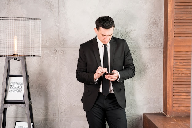 Foto gratuita joven empresario con teléfono inteligente de pie por la pared